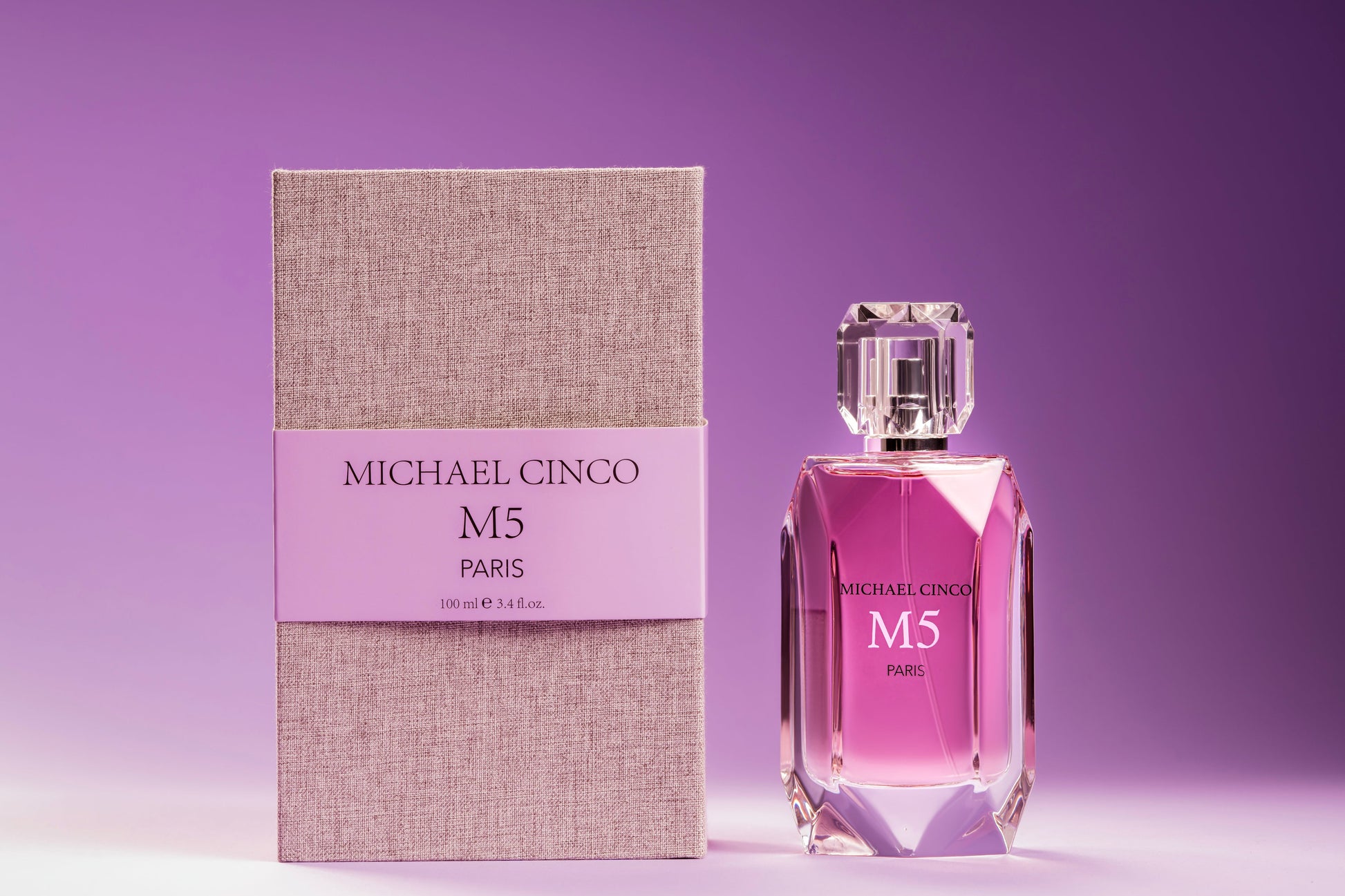 A bottle of Fragrapedia Haus M5 Eau de Parfum by Michael Cinco stands next to its textured beige and lavender box, both set against a gradient purple background. The faceted bottle with a clear cap suggests the seductive fragrance inside.