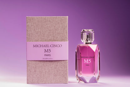 A bottle of Fragrapedia Haus M5 Eau de Parfum by Michael Cinco stands next to its textured beige and lavender box, both set against a gradient purple background. The faceted bottle with a clear cap suggests the seductive fragrance inside.