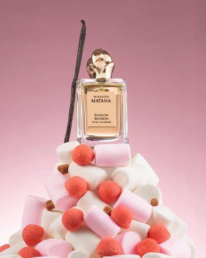 The Evasion Bonbon Extrait de Parfum by Maison Mataha rests on a bed of pink and white marshmallows, red candy, and a vanilla bean, evoking a cozy candy dream against a pink gradient background.