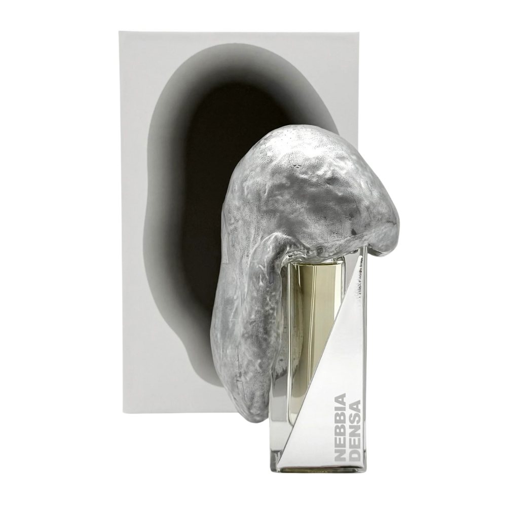 The NEBBIA DENSA "dense fog" Eau de Parfum by Filippo Sorcinelli comes in a bottle adorned with an abstract metallic silver form. A white panel behind it, cast with a dark shadow, hints at sandalwood and botanical moss notes.