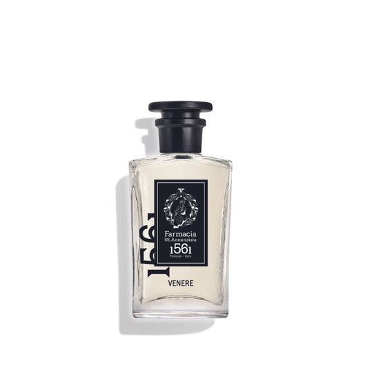 A clear glass bottle of Venere Eau de Parfum by Farmacia SS. Annunziata 1561, featuring a black cap, rests against a white background. Its soft shadow hints at the alluring amber and musk notes inside.