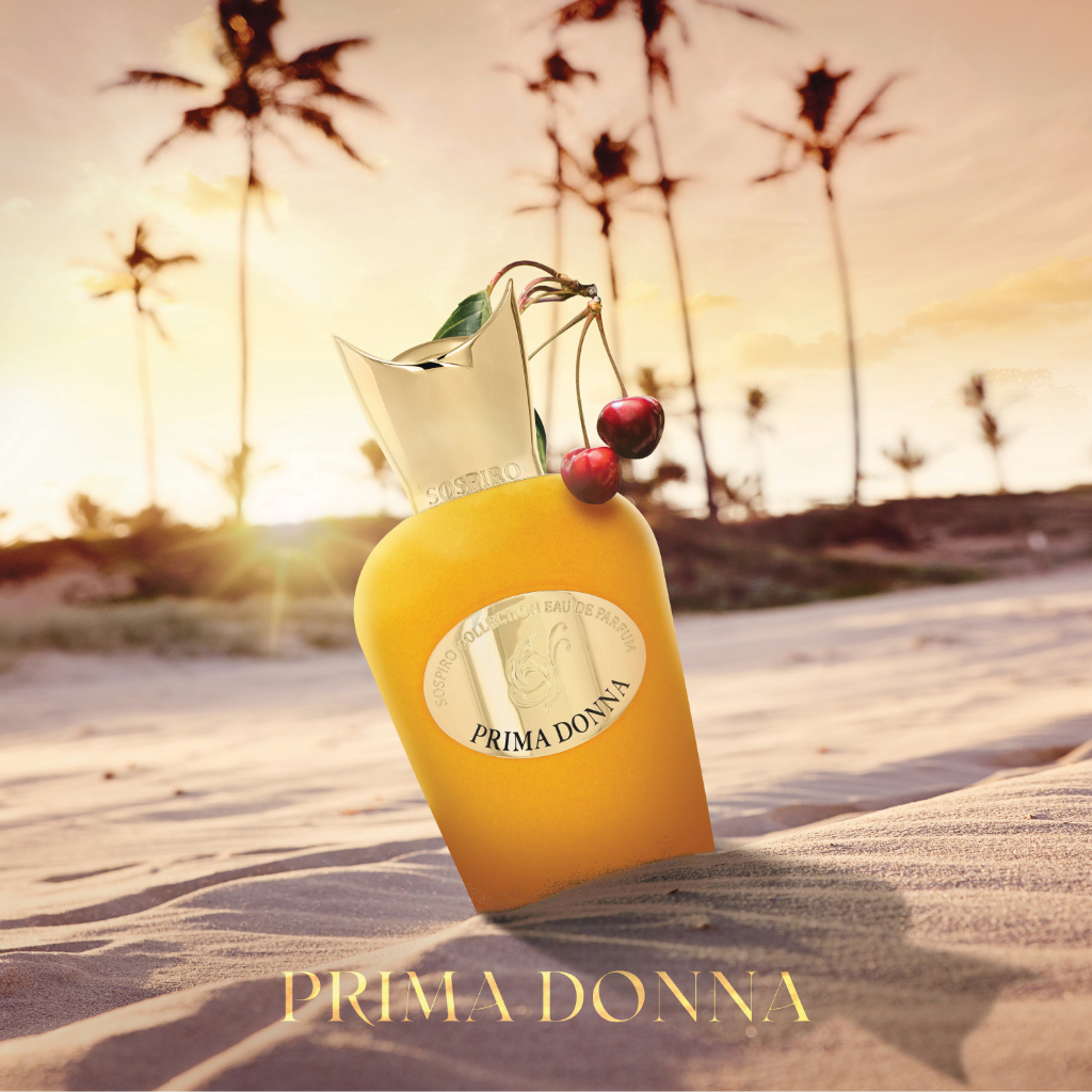A golden Prima Donna Eau de Parfum by SOSPIRO, adorned with cherries and a leaf on its cap, rests on sandy ground against palm trees and a sunset. This floral fragrance shines with "PRIMA DONNA" at the base.