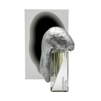 The NEBBIA FITTA "thick fog" Eau de Parfum by Filippo Sorcinelli is a clear glass bottle, partially veiled by a metallic silver sculpture. Oakmoss notes are suggested, while a white background and gray shadow lend modern elegance.