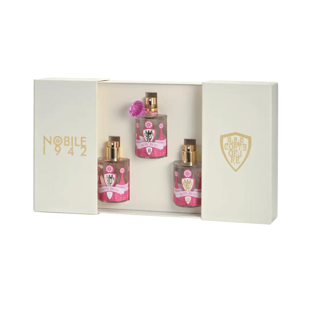 A cream-colored BON BON - Nobile 1942 gift box by Nobile 1942 with gold logos and sliding panels holds three fairy tale fragrances in pink-labeled bottles, beautifully arranged in a row.