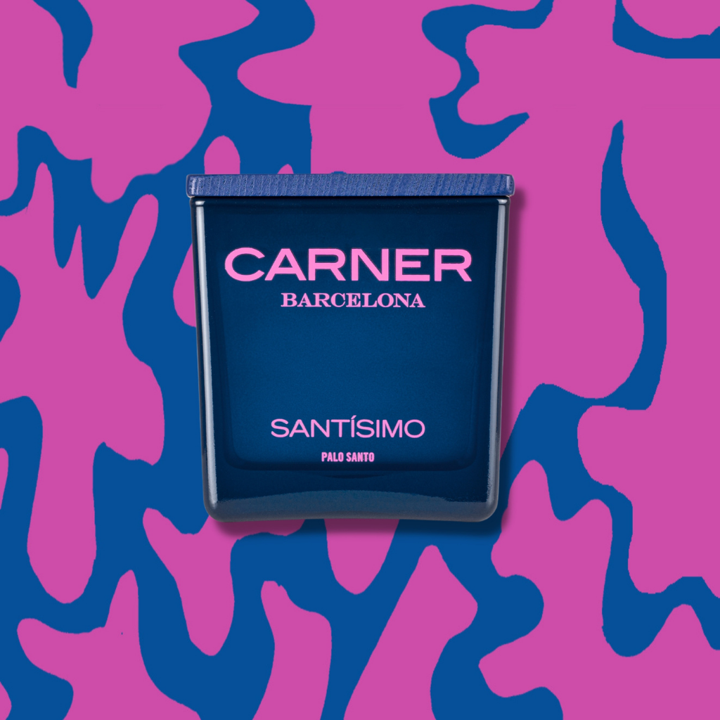 A dark blue SANTÍSIMO CANDLE by Carner is centered on a pink and blue abstract patterned background. The candle label features pink and white text.