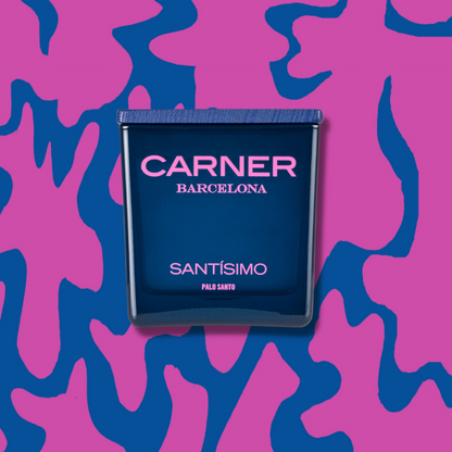 A dark blue SANTÍSIMO CANDLE by Carner is centered on a pink and blue abstract patterned background. The candle label features pink and white text.