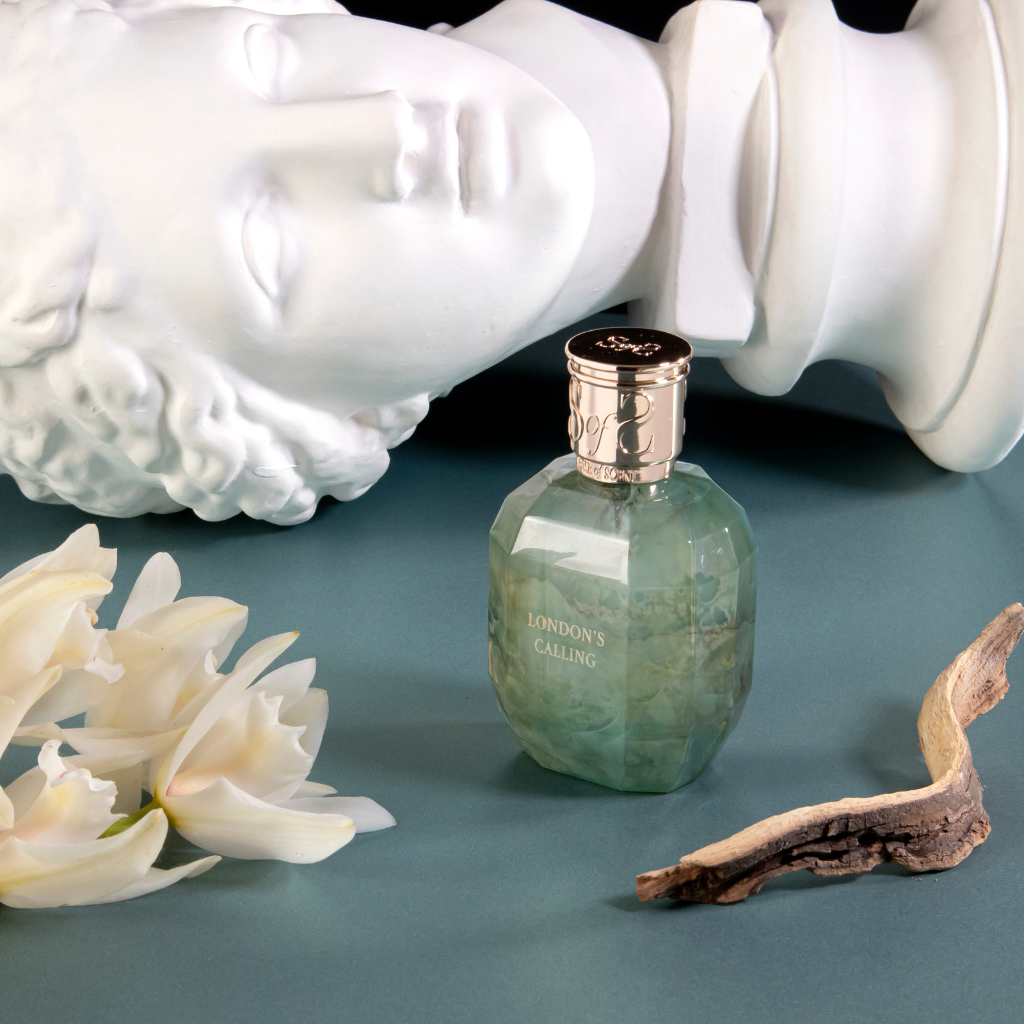 The LONDON'S CALLING Eau de Parfum by Sense of Scent rests on a blue surface with white flowers, driftwood, and a classical white face sculpture lying nearby.