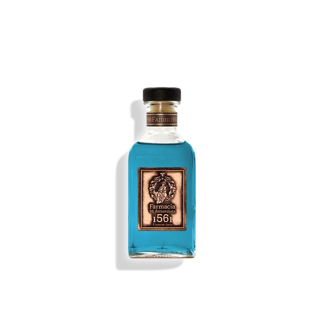 A clear glass bottle with a black cap holds bright blue liquid. The ornate label reads “Farmacia SS. Annunziata 1561.” The Fabbri DIFFUSER is perfect as a home fragrance diffuser with black sticks, casting a shadow on a white background.