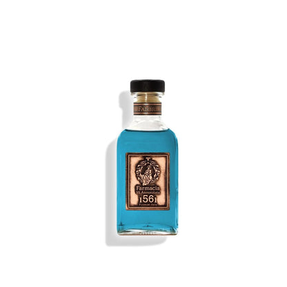 A clear glass bottle with a black cap holds bright blue liquid. The ornate label reads “Farmacia SS. Annunziata 1561.” The Fabbri DIFFUSER is perfect as a home fragrance diffuser with black sticks, casting a shadow on a white background.