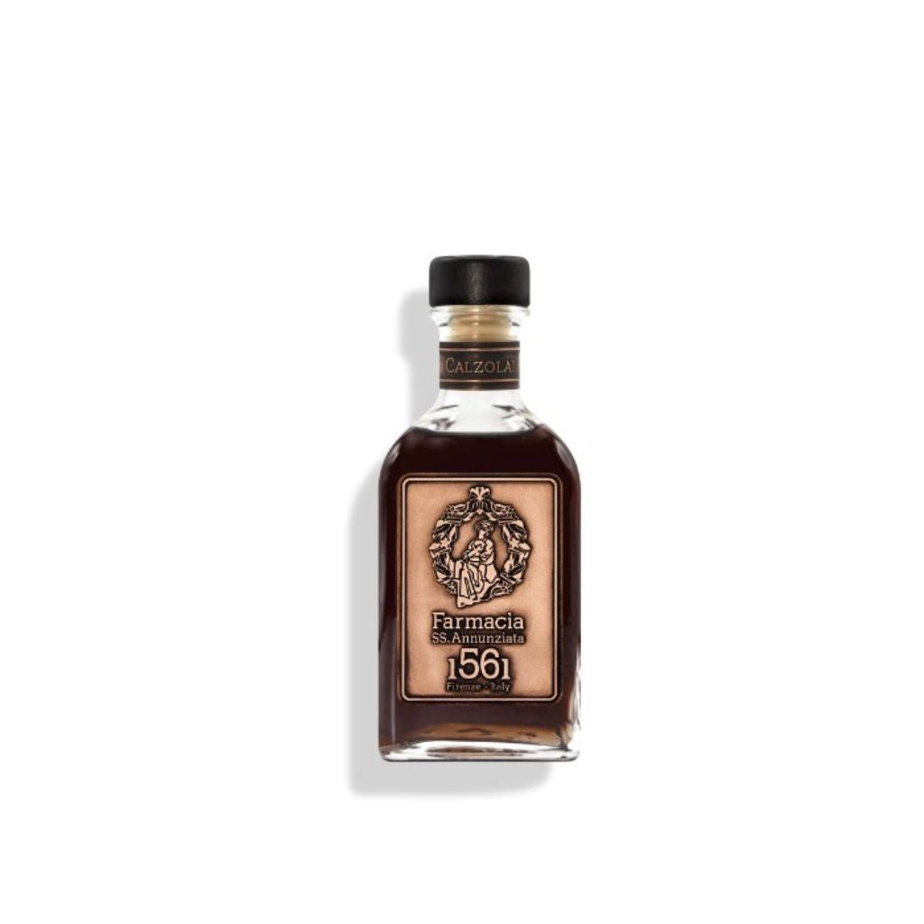 A glass Calzolai DIFFUSER by Farmacia SS. Annunziata 1561, with a black cap and ornate emblem, contains dark liquid and is photographed against a white background. It is often used as a home fragrance leather diffuser.