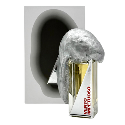 The VENTO IMPETUOSO "impetuous wind" Eau de Parfum by Filippo Sorcinelli, 50 ml, features a sculptural silver cap and sits before an abstract background with a metallic form. Sophisticated vetiver notes add elegance.
