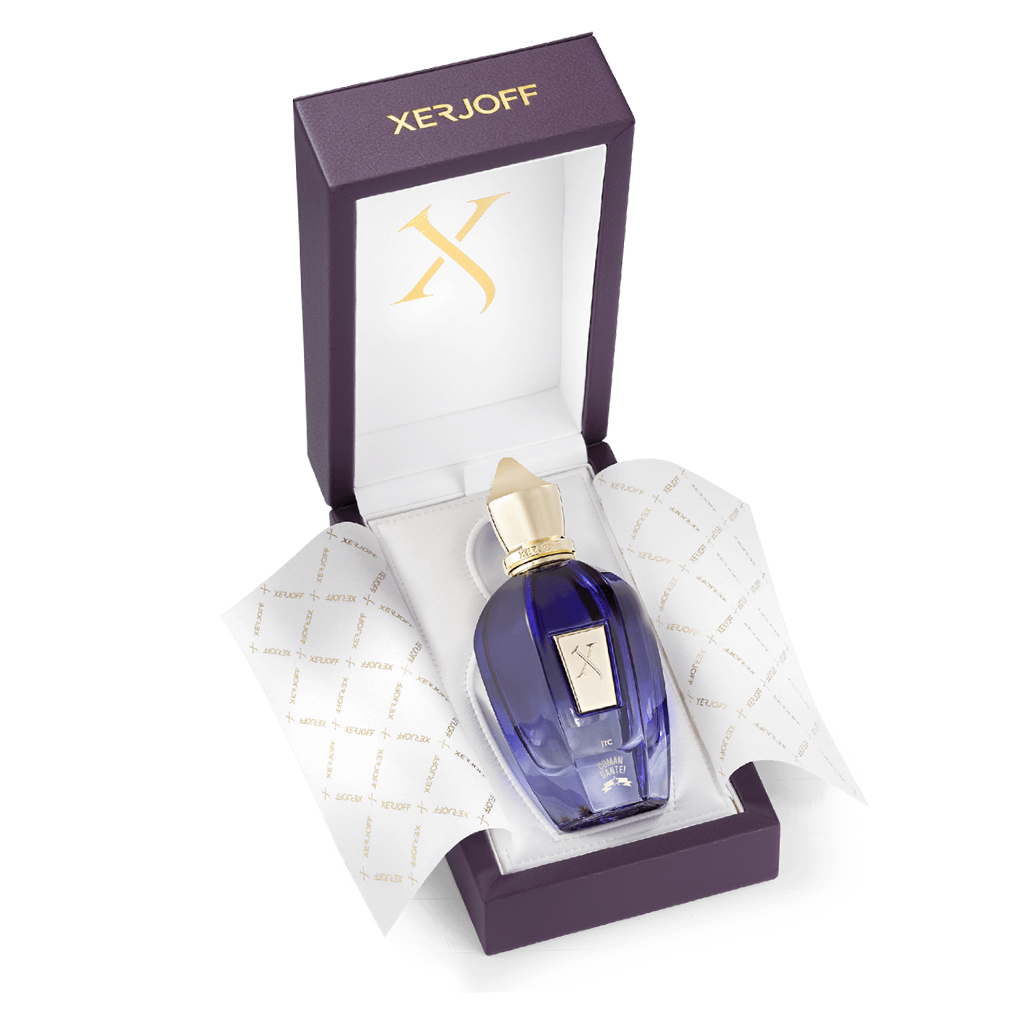 A blue Comandante Eau de Parfum by XERJOFF with a gold cap sits in an open purple box lined with branded white tissue and gold logo accents, hinting at the elegant tobacco scent inside.
