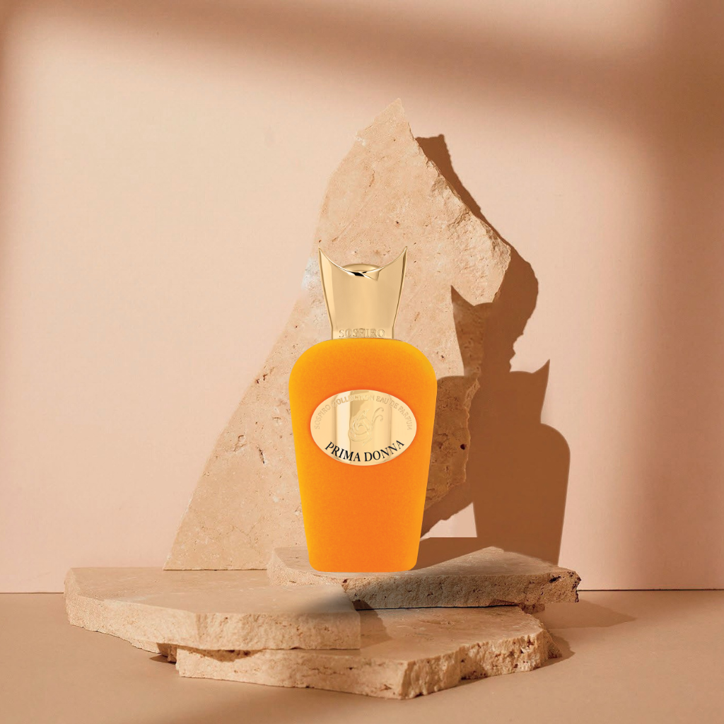 A vibrant orange Prima Donna Eau de Parfum by SOSPIRO with a gold cap rests on stone slabs against a beige background, highlighted by dramatic light and shadow to reflect its captivating floral scent.