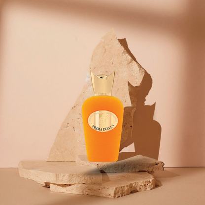 A vibrant orange Prima Donna Eau de Parfum by SOSPIRO with a gold cap rests on stone slabs against a beige background, highlighted by dramatic light and shadow to reflect its captivating floral scent.
