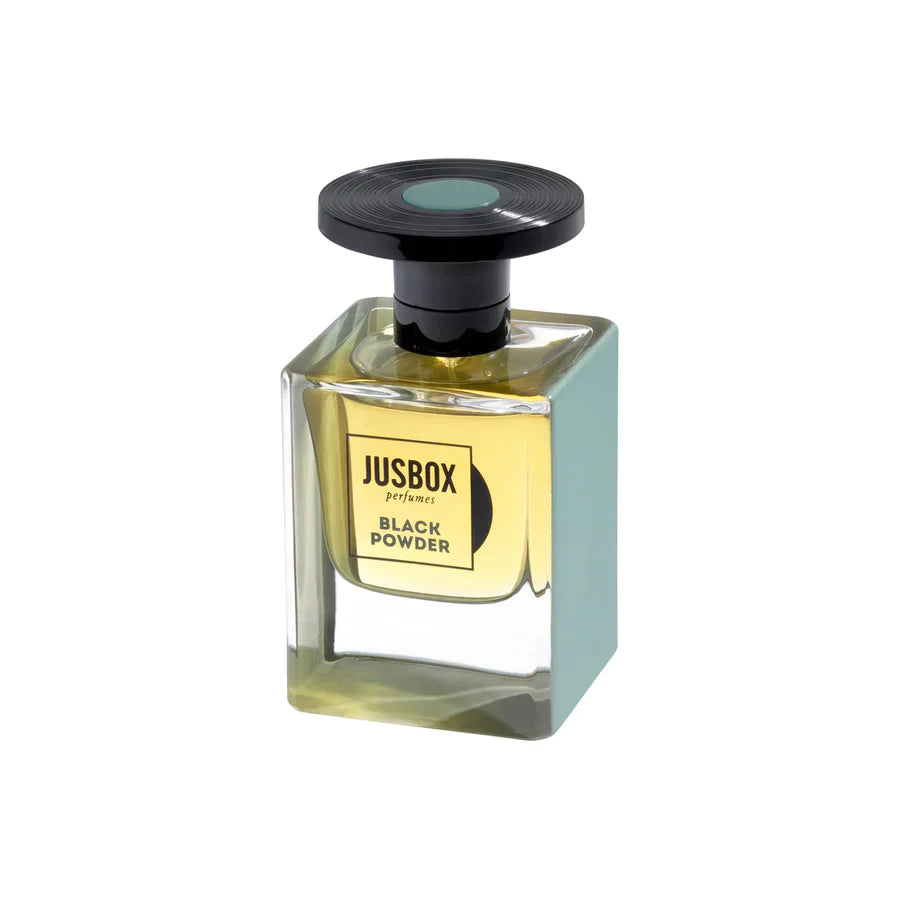 A rectangular bottle of Jusbox Black Powder Eau de Parfum, a rebellious leather fragrance, contains yellow liquid and is crowned with a large round black cap. The front label shows the Jusbox brand and perfume name.