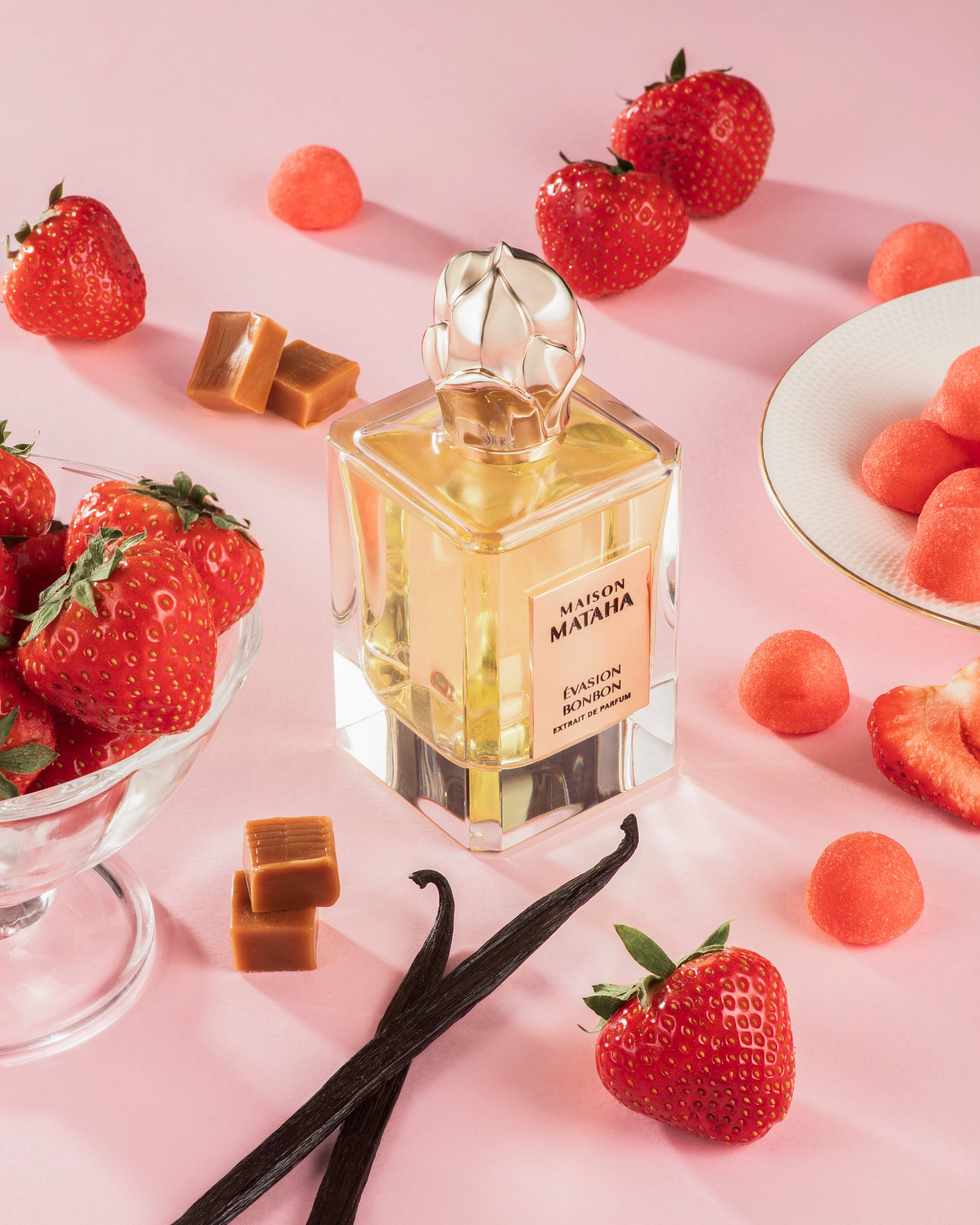 A bottle of Evasion Bonbon Extrait de Parfum by Maison Mataha rests on a pink surface, surrounded by fresh strawberries, caramel candies, vanilla beans, a bowl of strawberries, and a plate with candy drops.