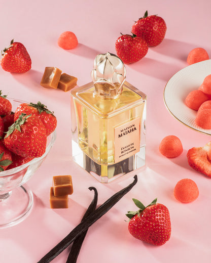 A bottle of Evasion Bonbon Extrait de Parfum by Maison Mataha rests on a pink surface, surrounded by fresh strawberries, caramel candies, vanilla beans, a bowl of strawberries, and a plate with candy drops.