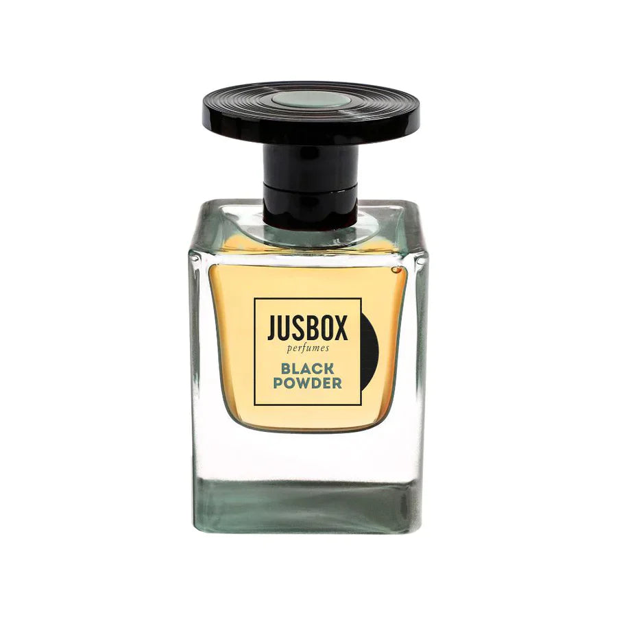A clear, square glass bottle of Jusbox Black Powder Eau de Parfum features a black round cap and light gold label, filled with amber-hued liquid—a bold leather accord perfume with a rebellious scent.