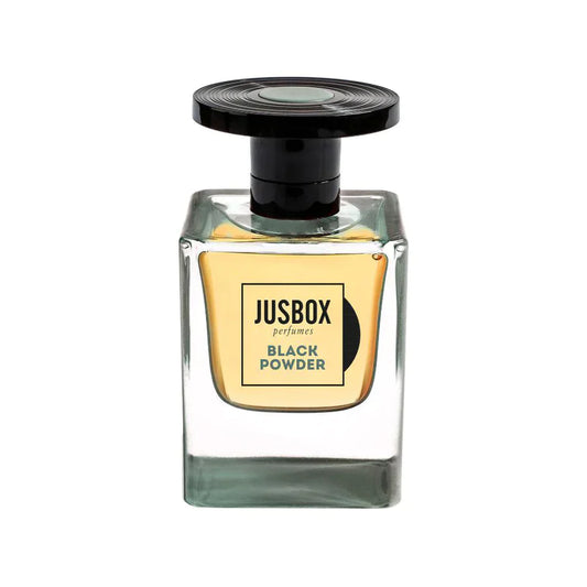 A clear, square glass bottle of Jusbox Black Powder Eau de Parfum features a black round cap and light gold label, filled with amber-hued liquid—a bold leather accord perfume with a rebellious scent.