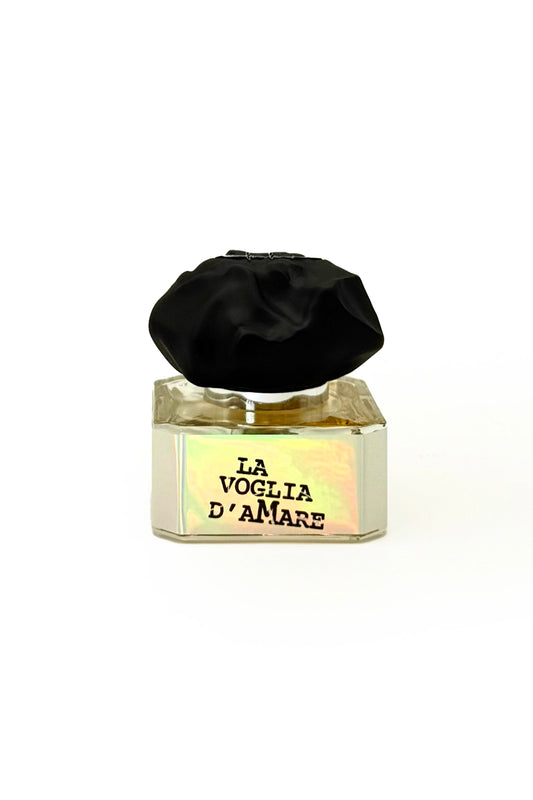 A square perfume bottle with a large black cap and gold label reading LA VOGLIA D'AMARE, the Eau de Parfum by Filippo Sorcinelli, evoking summer nights, displayed against a plain white background.
