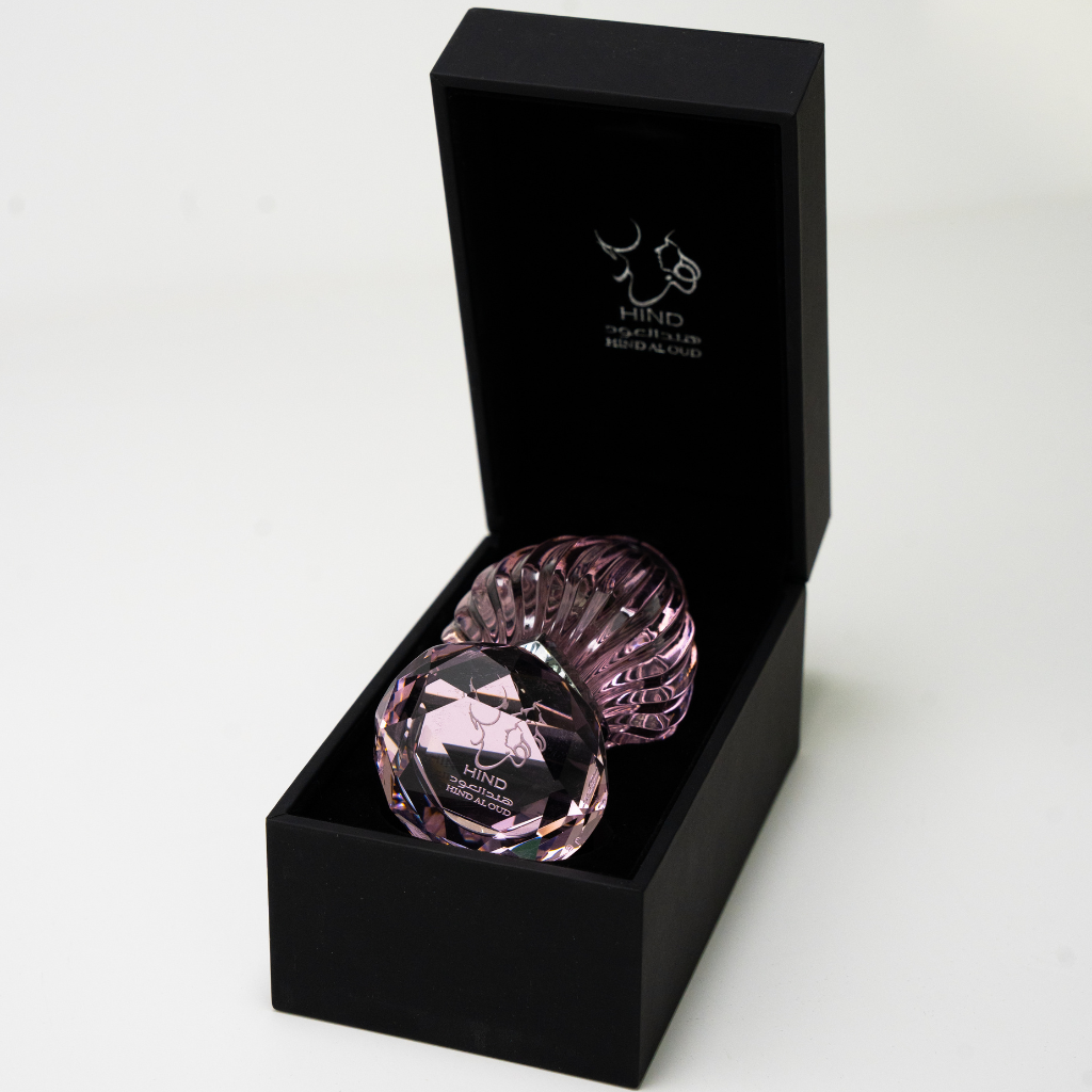 A pink, faceted glass bottle of Hind Al Oud's Pink Oud Concentrated Oil 6ml, with "Shiekh Zayed Road - Dubai" engraved on the lid, sits in a black box. Notes of agarwood and jasmine hint at its luxurious fragrance.