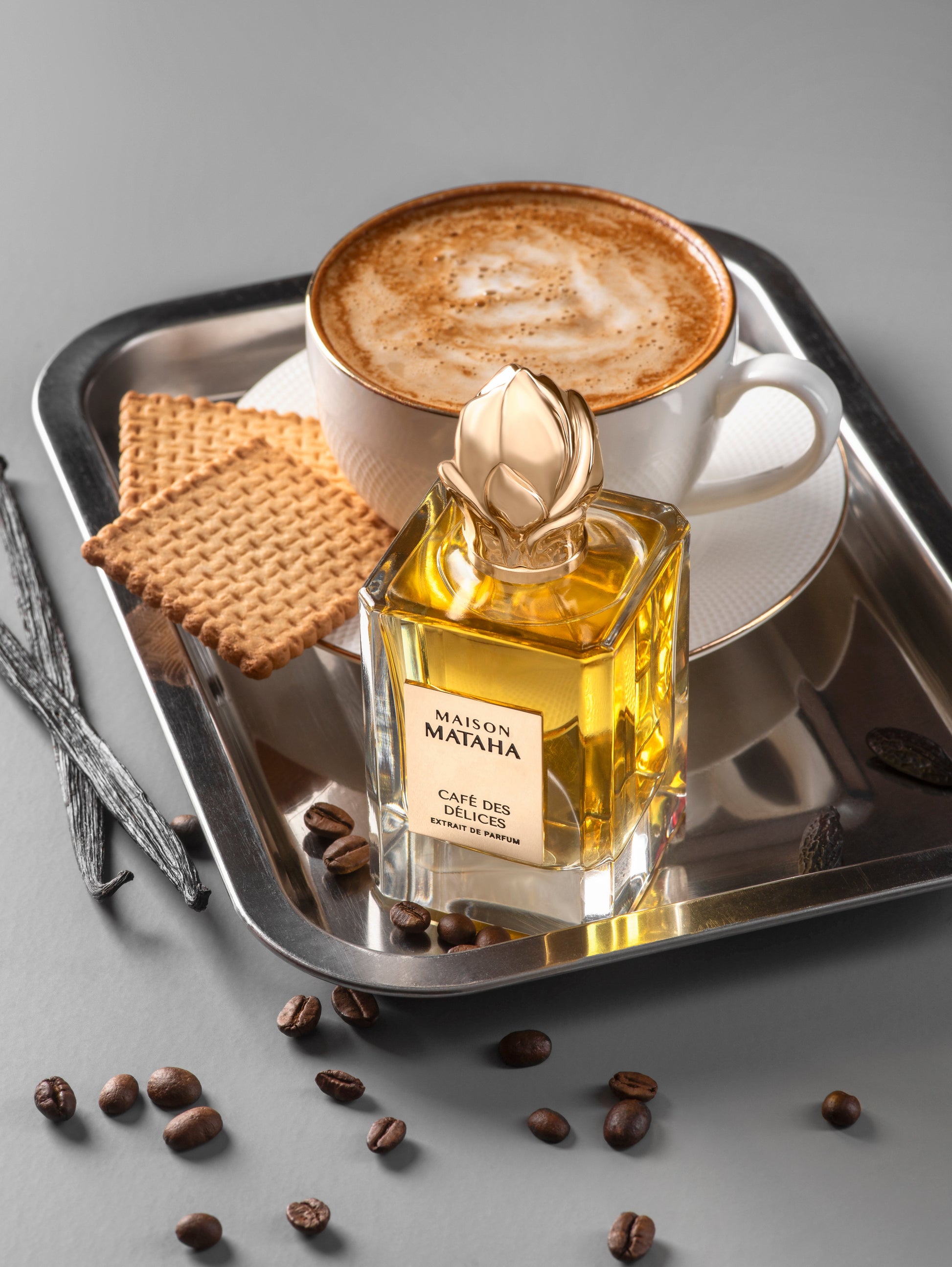 A cup of cappuccino and two rectangular biscuits rest on a silver tray beside Maison Mataha’s Café des Délices Extrait de Parfum, surrounded by coffee beans and vanilla pods on a grey surface, evoking rich coffee notes.