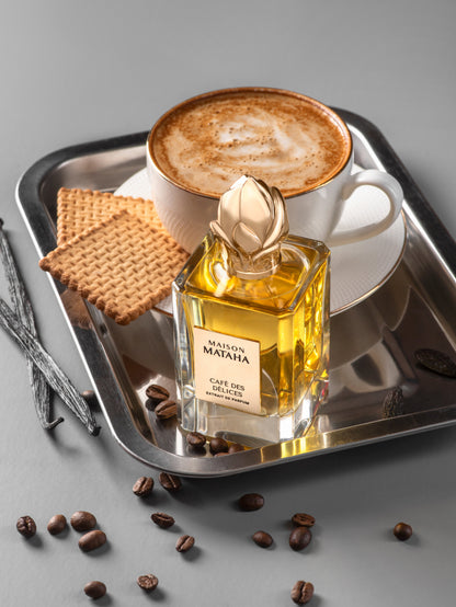 A cup of cappuccino and two rectangular biscuits rest on a silver tray beside Maison Mataha’s Café des Délices Extrait de Parfum, surrounded by coffee beans and vanilla pods on a grey surface, evoking rich coffee notes.