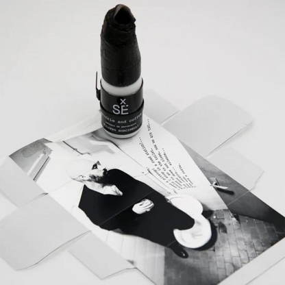 A black-and-white photo of a seated, white-bearded man rests on a white box. On top is Filippo Sorcinelli’s X SÉ QUICKIE AND COFFEE Eau de Parfum, its wax-sealed bottle exuding an intense vanilla and coffee scent.