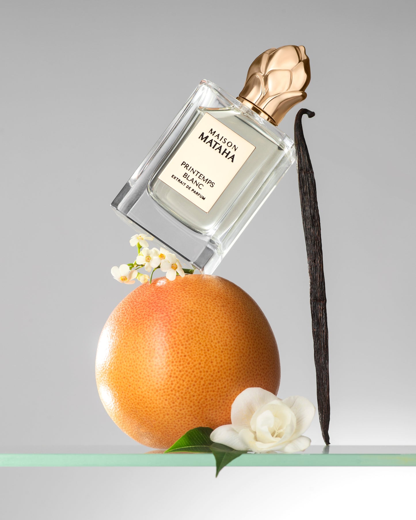 A bottle of Printemps Blanc Extrait de Parfum by Maison Mataha, celebrated for its floral notes and jasmine sambac, is balanced on a grapefruit with a vanilla bean, white flowers, and green leaves set against a light gray background.