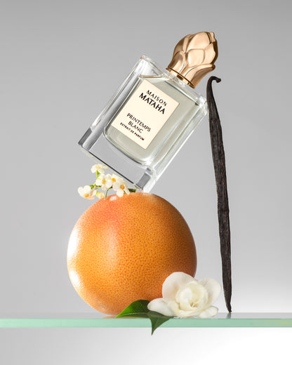 A bottle of Printemps Blanc Extrait de Parfum by Maison Mataha, celebrated for its floral notes and jasmine sambac, is balanced on a grapefruit with a vanilla bean, white flowers, and green leaves set against a light gray background.