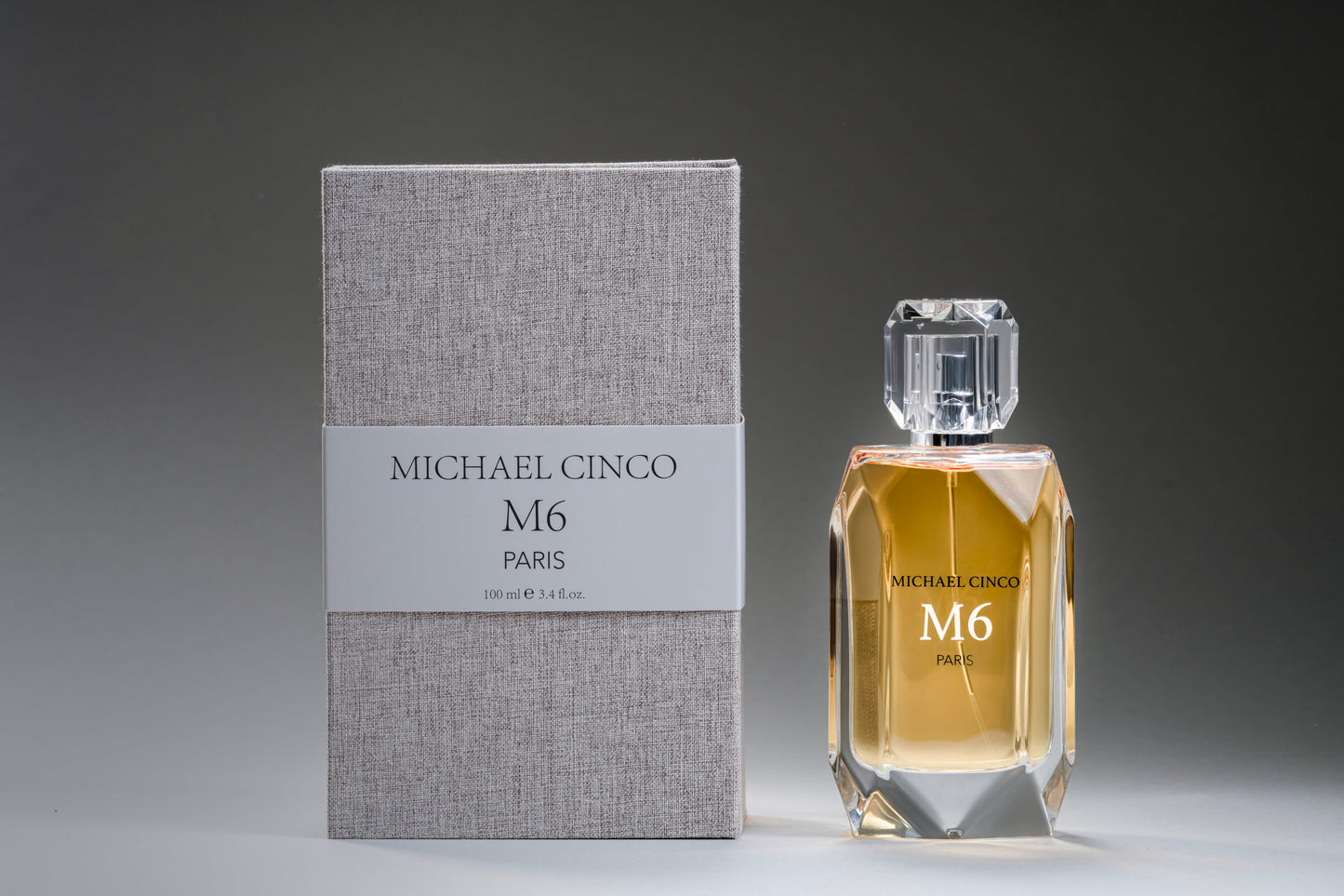 A bottle of M6 Eau de Parfum by Fragrapedia Haus sits beside a textured grey box, featuring a clear faceted cap and an elegant design that evokes the allure of patchouli.