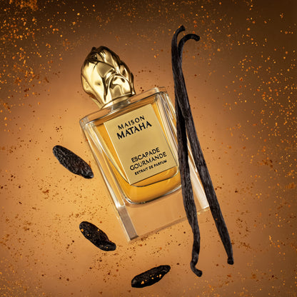 The Escapade Gourmande Extrait de Parfum by Maison Mataha, featuring a golden cap, is displayed among vanilla and tonka beans, radiating a decadent crème brûlée scent on an amber background with sparkling accents.