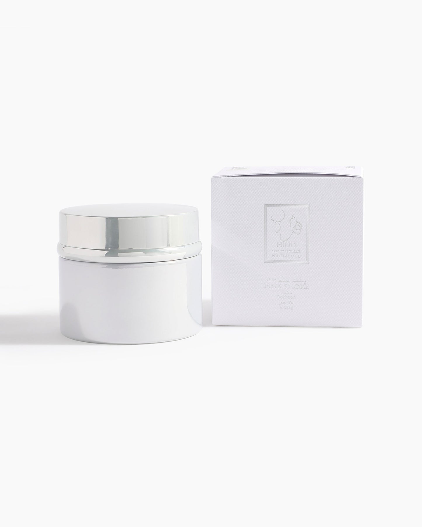 A white cosmetic jar with a shiny silver lid sits beside a matching white box with Hind Al Oud branding, set against a plain backdrop—showcasing Pink Smoke Dokhoon by Hind Al Oud, inspired by jasmine and pink oud.