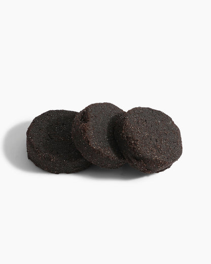 Three round, dark chocolate cookies are stacked on a plain white background with a soft shadow—an inviting scene reminiscent of the rich, alluring notes found in Hind Al Oud's Pink Smoke Dokhoon fragrance.