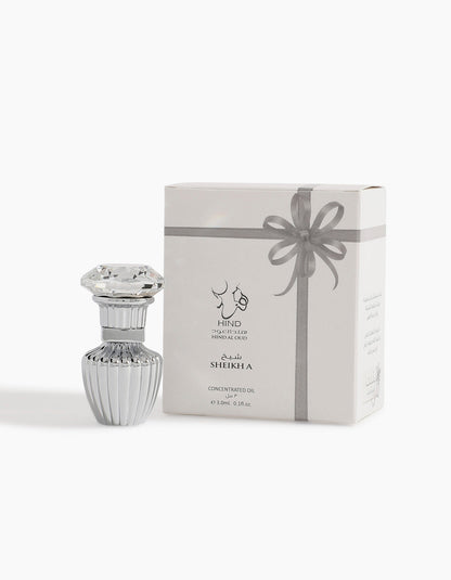 A decorative glass bottle with a gem-like lid stands next to a white box featuring silver Arabic and English text and a gray ribbon. Sheikh A Petite Concentrated Oil 3ml by Hind Al Oud exudes luxury with rare ambergris notes.