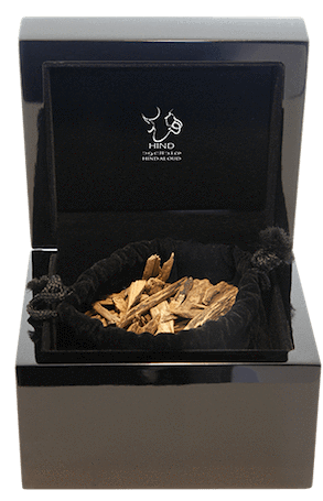 A black, glossy box with a hinged lid opens to reveal Oud Al Mezayeen Natural Oud chips in a velvet pouch. The inside lid displays Hind Al Oud’s logo and white text, reflecting the brand’s luxurious essence.
