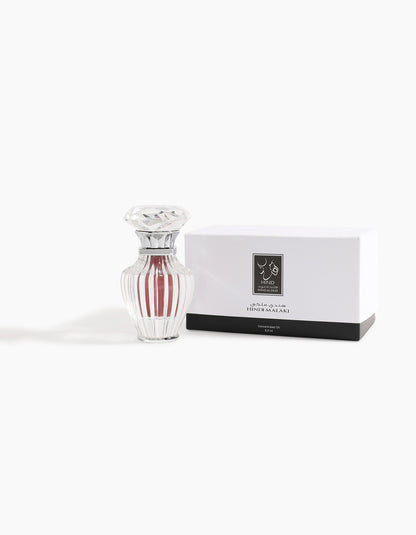 A clear, ribbed glass bottle with a faceted lid sits beside a white box in black and silver, both labeled in Arabic and English. The box displays Hind Al Oud's Hindi Malaki Pure Dehn Oud Concentrated Oil 6ml—a refined agarwood scent.
