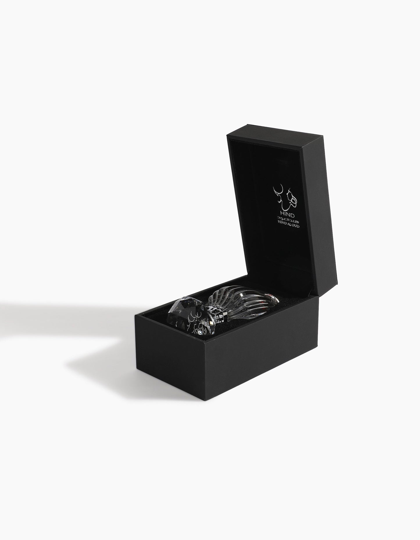 A black rectangular box with a hinged lid, partially open to reveal black utensils inside. The inside lid features the Hind Al Oud logo and text, highlighting the elegance of Hindi Malaki Pure Dehn Oud Concentrated Oil 6ml. The box casts a shadow on white.