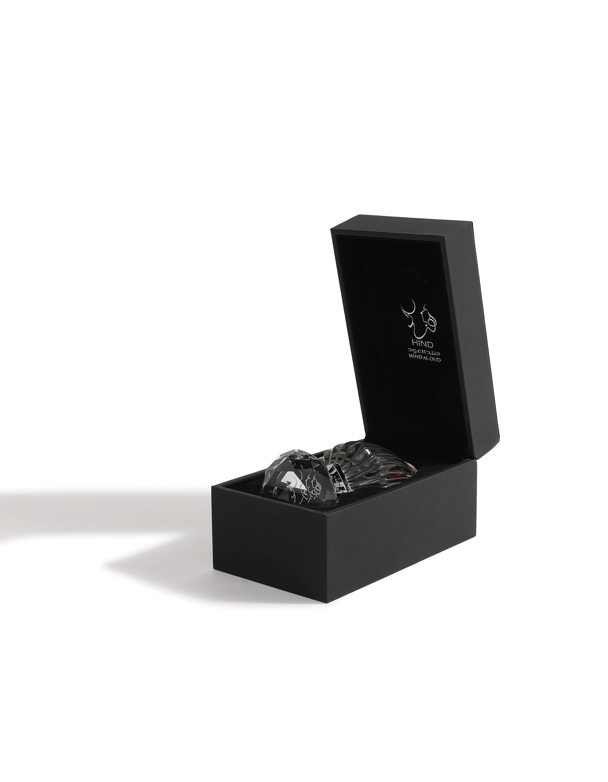 A black rectangular box with the lid open reveals a clear glass bottle of Sheikh A Jewel Concentrated Oil 6ml by Hind Al Oud. Inside the lid is a simple white graphic and brand text. The box casts a shadow on a white surface.