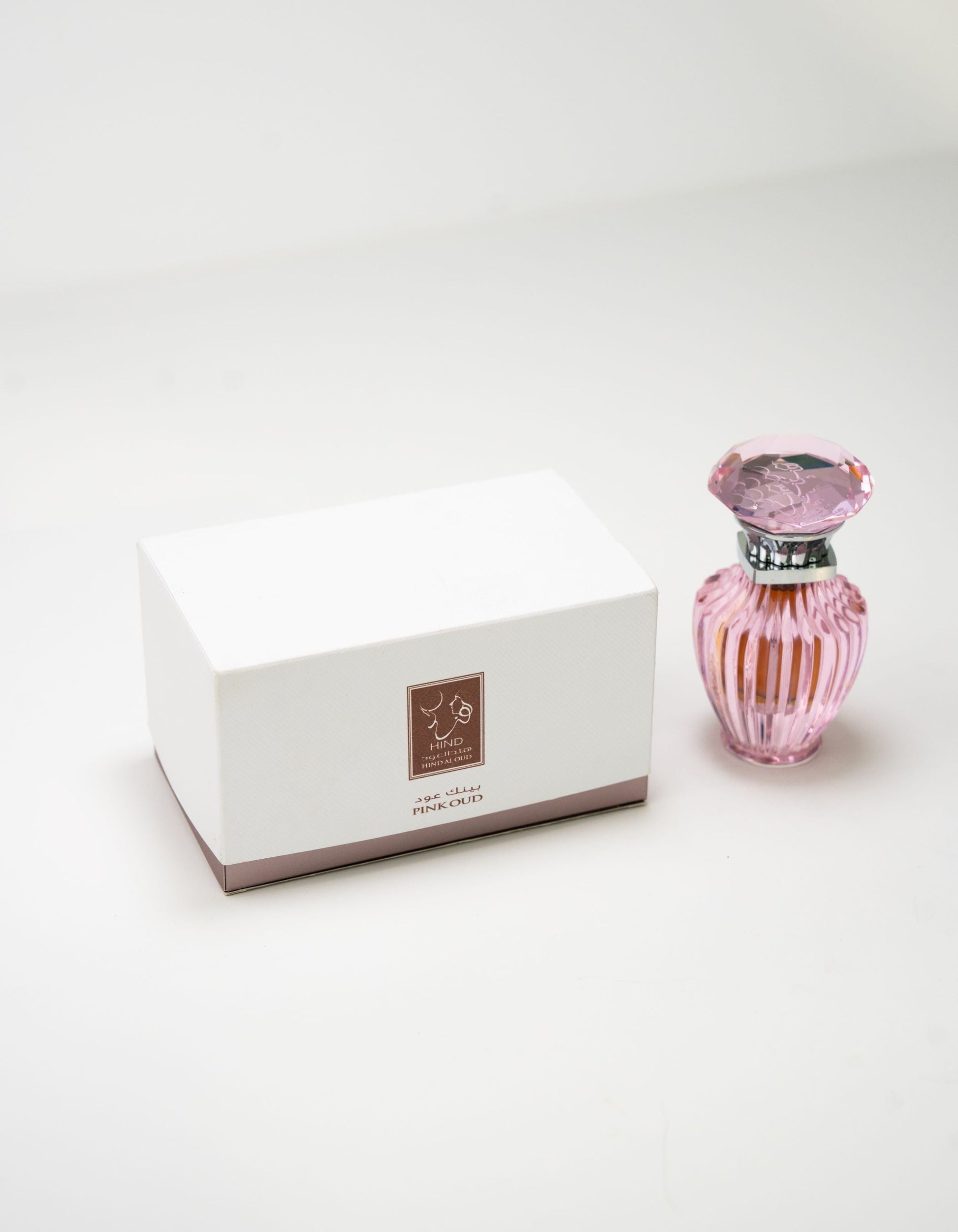 A white rectangular box with a brown label stands beside Hind Al Oud’s Pink Oud Concentrated Oil 6ml—a pink, faceted bottle with a decorative cap—evoking elegant notes of jasmine and cashmere wood on a plain white surface.