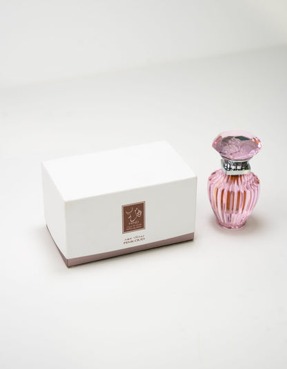 A white rectangular box with a brown label stands beside Hind Al Oud’s Pink Oud Concentrated Oil 6ml—a pink, faceted bottle with a decorative cap—evoking elegant notes of jasmine and cashmere wood on a plain white surface.