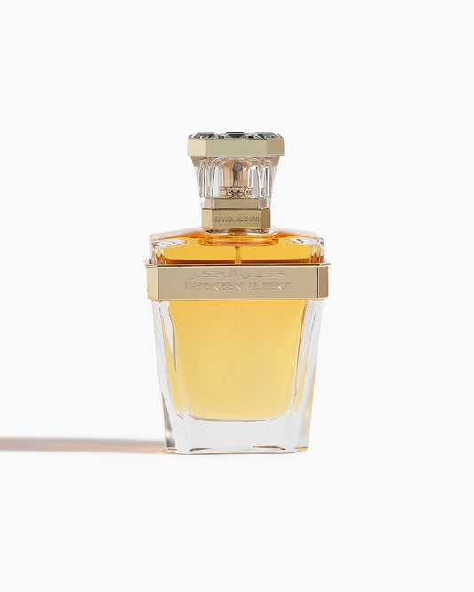A clear glass bottle of Kheneen Al Fekr Parfum by Hind Al Oud, with golden-yellow liquid and a gold cap, is labeled in gold and stands against a plain white background—evoking notes of cherry blossom and rich agarwood.