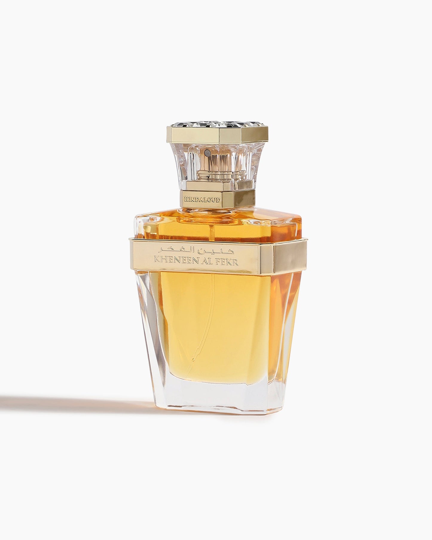 A clear, faceted glass perfume bottle with golden-yellow liquid and a gold band and cap, labeled Kheneen Al Fekr Parfum by Hind Al Oud—an elegant fragrance suggesting cherry blossom notes.
