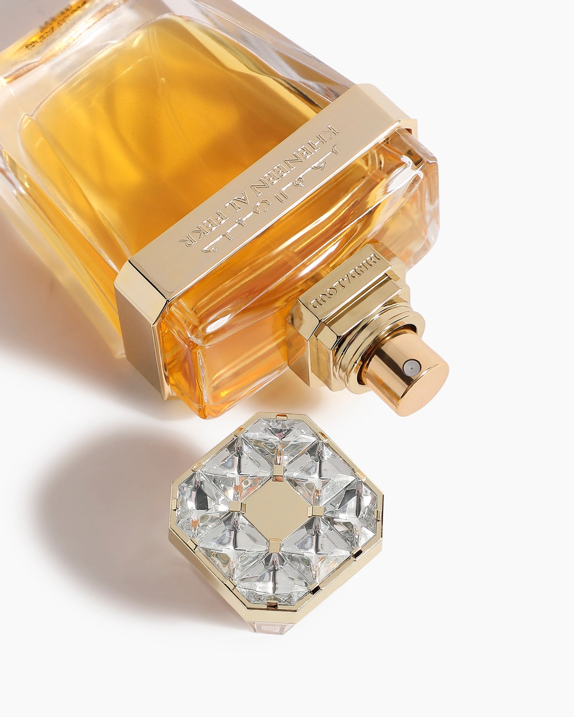 A close-up of Kheneen Al Fekr Parfum by Hind Al Oud shows a luxury bottle with golden accents and a gem-like cap beside it, set on a white background. The bottle is partially filled with amber-hued agarwood perfume.