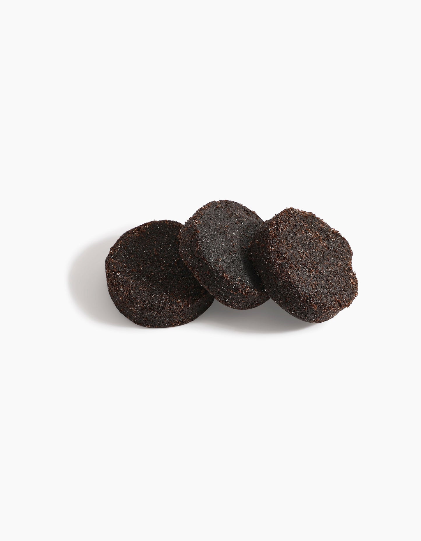 Three round, dark brown Hind Dokhoon - Hind Al Oud pucks by Hind Al Oud are arranged on a white background, with one puck leaning against the others and a soft shadow cast to the left.