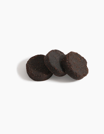 Three round, dark brown Hind Dokhoon - Hind Al Oud pucks by Hind Al Oud are arranged on a white background, with one puck leaning against the others and a soft shadow cast to the left.