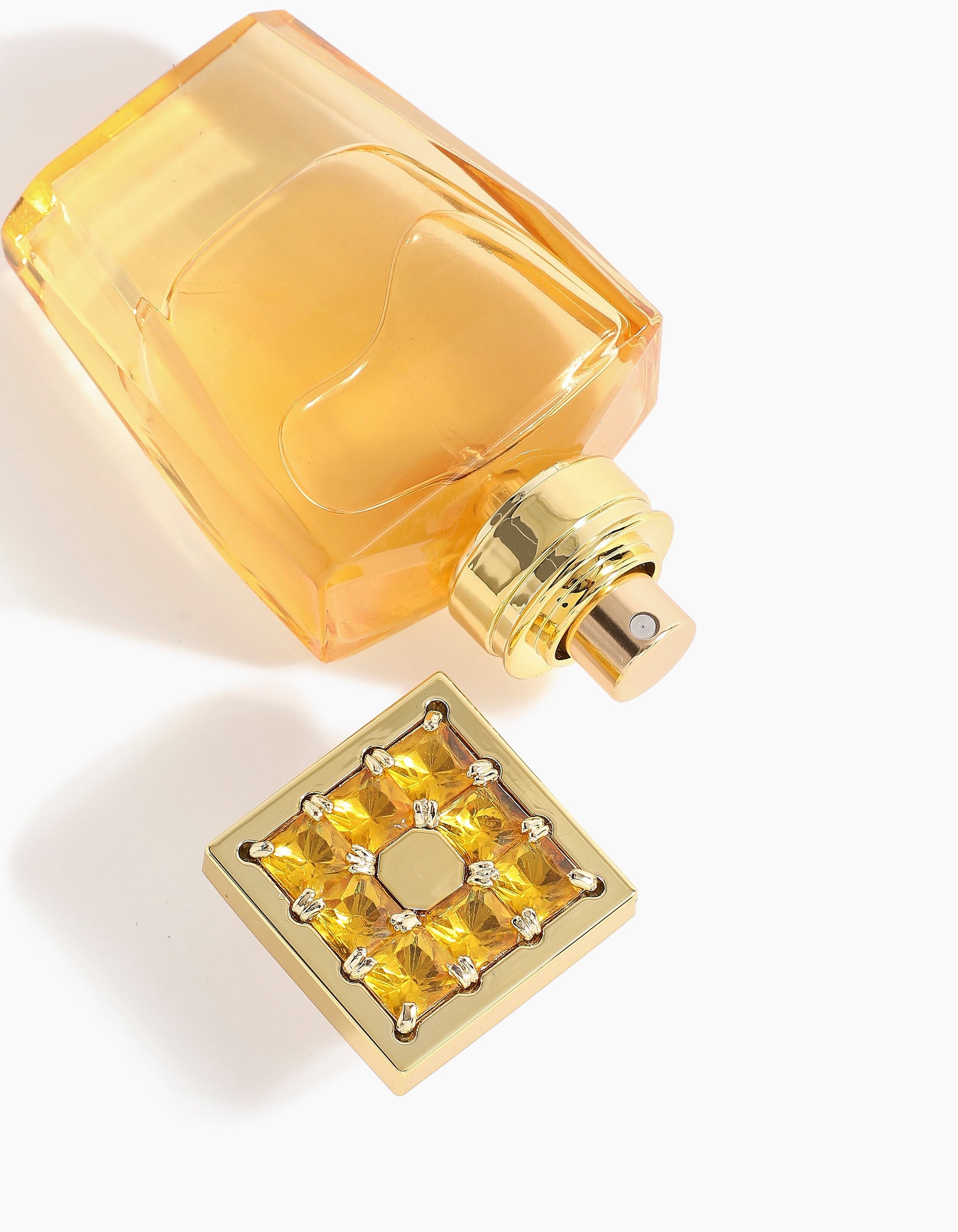 A golden Ahojas Hair Mist bottle by Hind Al Oud with a square, ornate cap embellished with yellow gemstones lies on its side against a white backdrop, cap off to reveal the spray nozzle and evoke its luxurious floriental aroma.