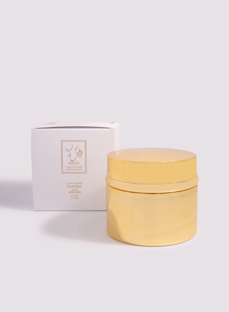 A shiny gold Ahojas Dokhoon - Hind Al Oud jar from Hind Al Oud, with a matching lid, stands before a white box with gold-foil details and text, evoking the fragrance’s luxurious fig and tobacco scent against a neutral backdrop.