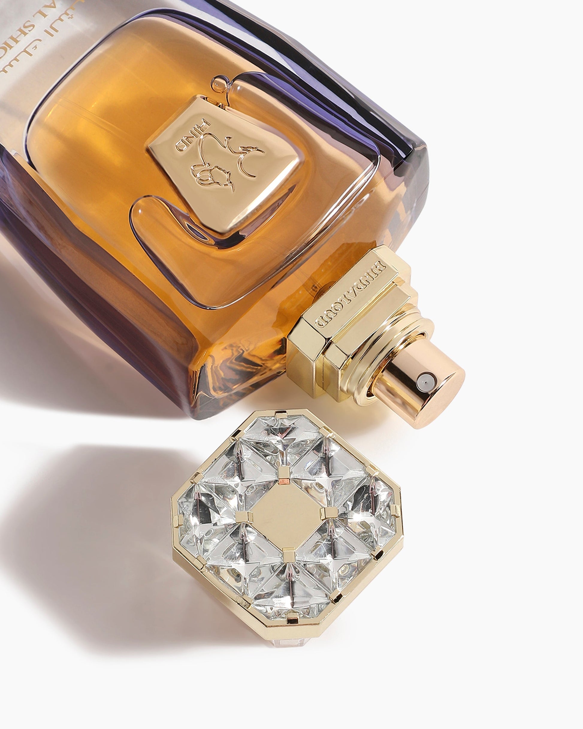 A close-up of the elegant Hind Al Oud Musk Al Shiokh Parfum bottle, tinted amber and purple, with a gold and clear gem-like cap beside it. The gold label features a camel and Arabic script.