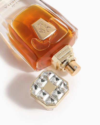 A close-up of the Hind Al Oud Sheikh A Parfum bottle with amber liquid and a gold, crystal-inspired geometric cap placed beside it on a white background.
